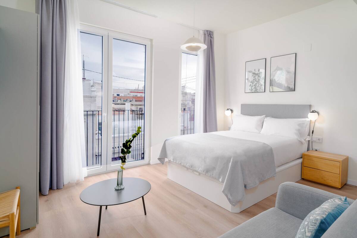 Un dormitorio moderno con una cama grande, mesitas de noche, una pequeña mesa redonda y vistas de la ciudad a través de amplias ventanas.