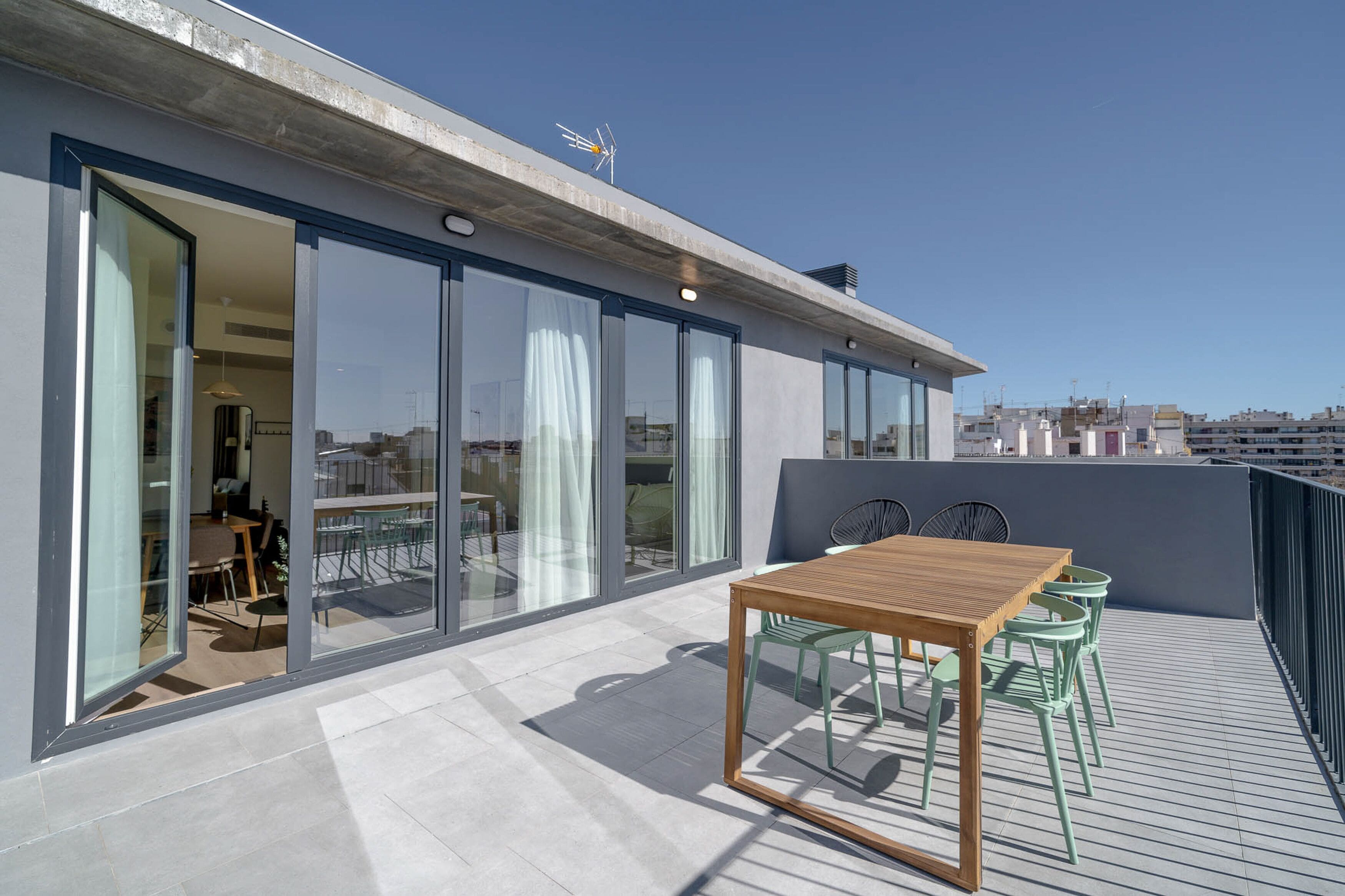 A rooftop terrace with a wooden table and chairs, large glass doors leading to an interior space, and a clear view of the city skyline.