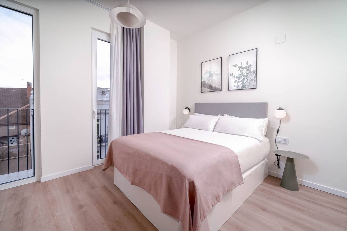 Un dormitorio con cama, una mesita de noche, lámpara, ventana con cortinas y dos cuadros en la pared.