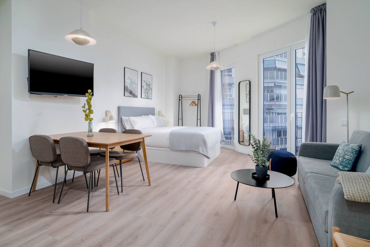 Una habitación de hotel moderna con cama, zona de comedor con sillas, mesita, sofá, televisor y un amplio ventanal con vista a edificios.