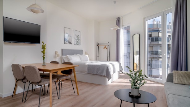 Una habitación de hotel moderna con una cama, una mesa de comedor con sillas, un televisor y un ventanal con vistas a la ciudad.