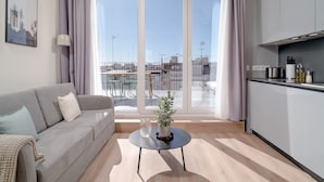 Una sala de estar moderna con un sofá gris, una mesita redonda con una planta en maceta y vistas de la ciudad a través de ventanas amplias.