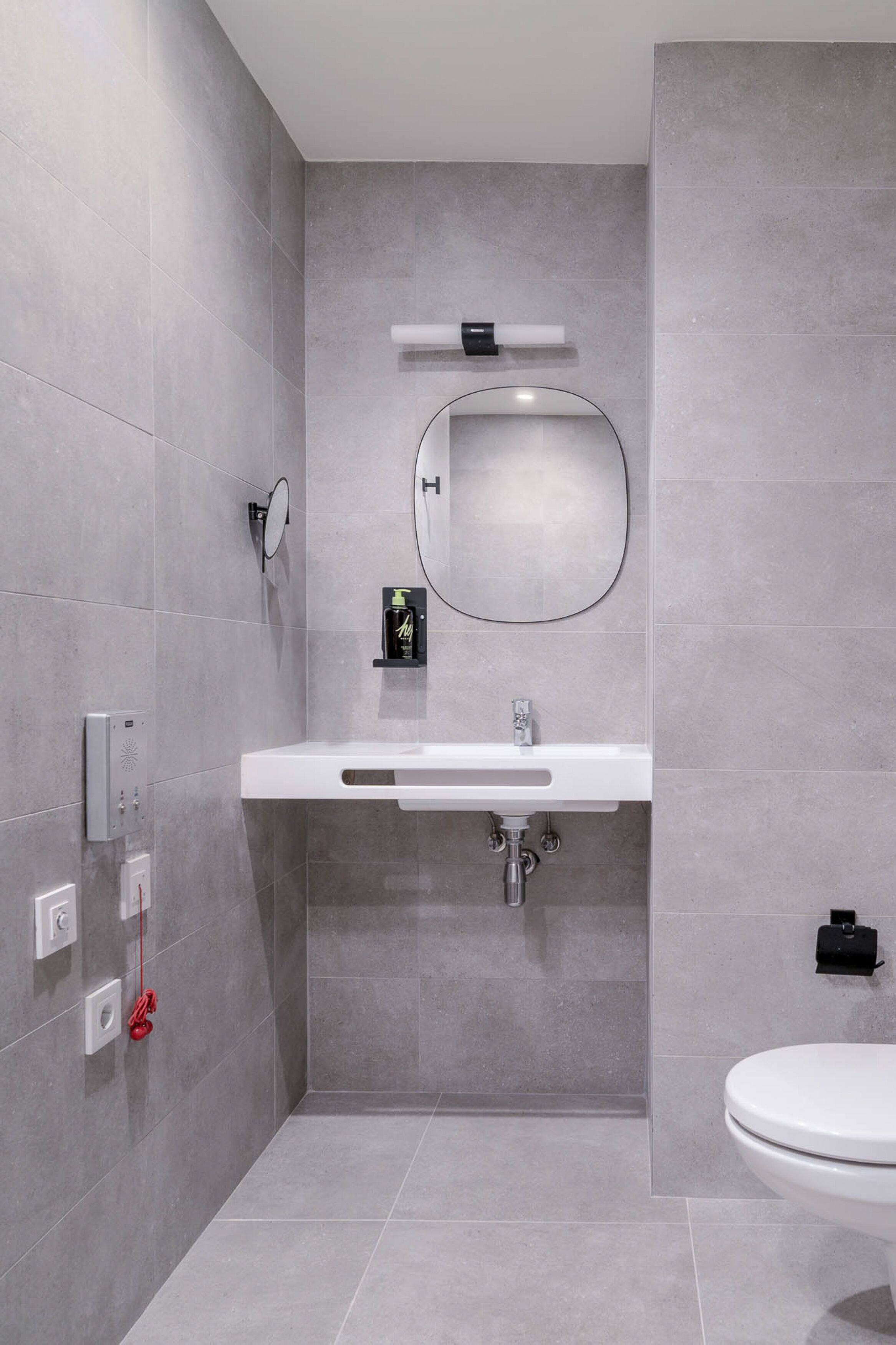 A modern bathroom with grey tiles, a white sink, a round mirror, and a toilet.