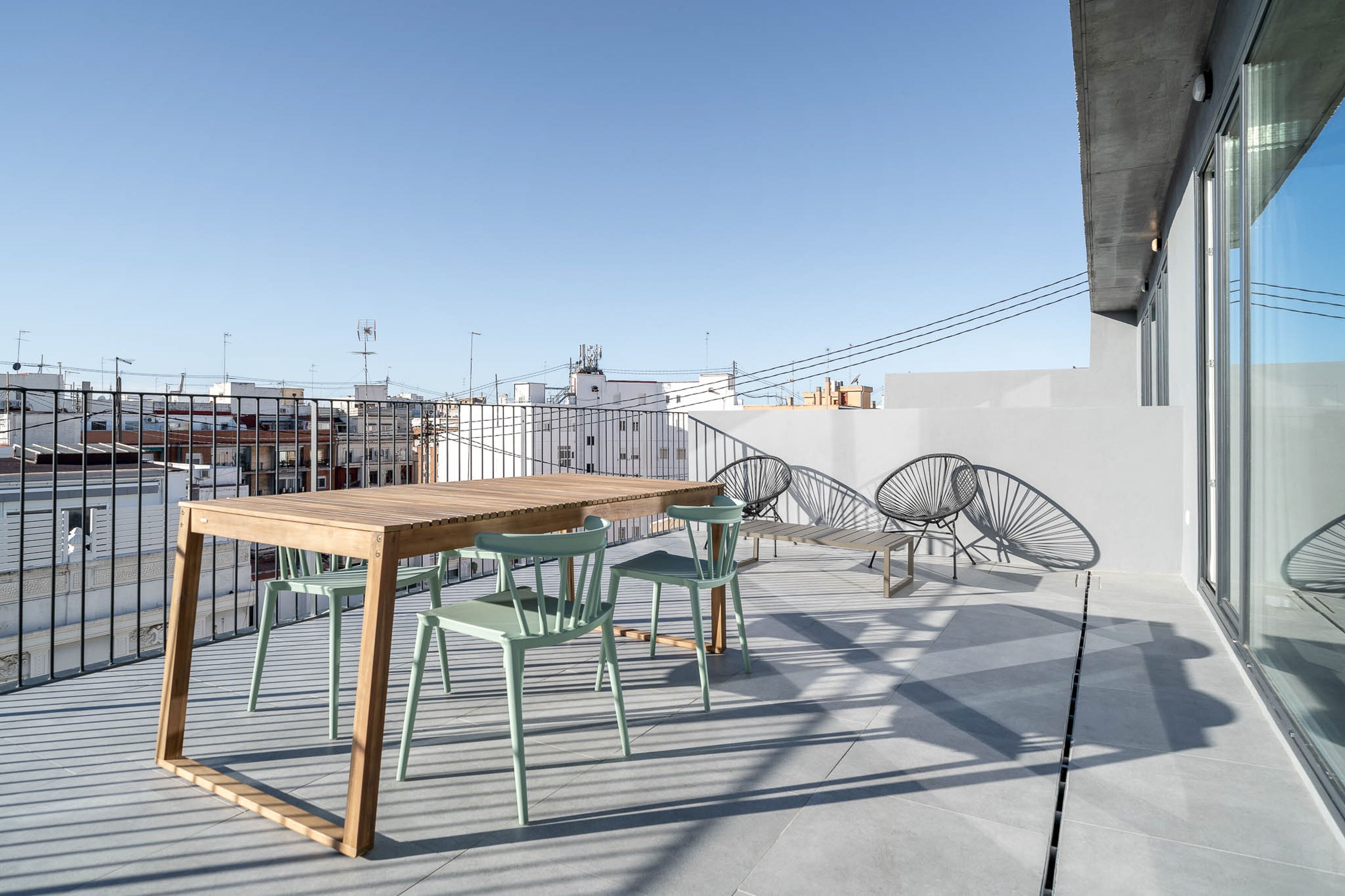 A rooftop terrace with a wooden table and green chairs, surrounded by a metal fence and overlooking a cityscape.