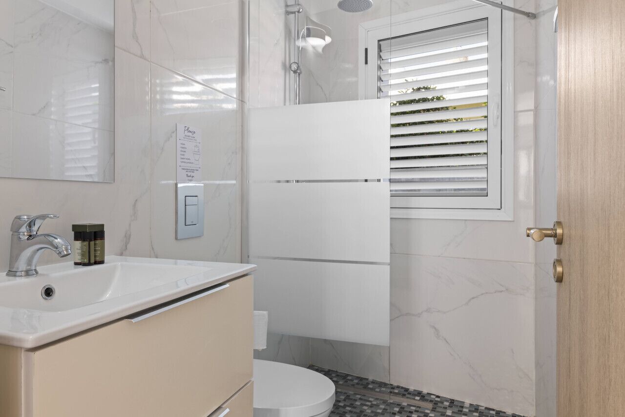 Un baño con lavamanos blanco, armarios color beige, un inodoro y una ducha con puerta de cristal.