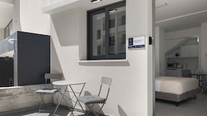 A modern hotel room with a bed, a small table, and a chair. There is a window with a view of the building's interior. - Phaedrus Living: Alkminis Seaside Suites Sand (Paphos)