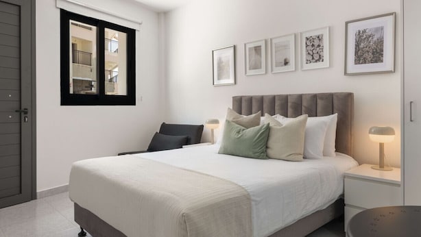 A bedroom with a bed, bedside tables, a window, and framed pictures on the wall. - Phaedrus Living: Alkminis Seaside Suites Sand (Paphos)