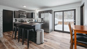 A modern kitchen with a central island, bar stools, a stainless steel refrigerator, and a view of a snowy outdoor area through a glass door. - Pet-friendly home with fenced-in yard and hot tub (Grawn)