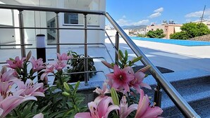 Un edificio moderno con fachada blanca y ventanas amplias. Frente al edificio, una escalera de metal con barandilla de vidrio. En primer plano, se plantan lirios rosados.