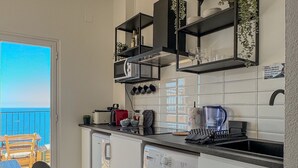 Une cuisine compacte dotée d’un lave-linge intégré, un coin repas avec une table et des chaises, et une vue sur la mer par une porte ouverte.