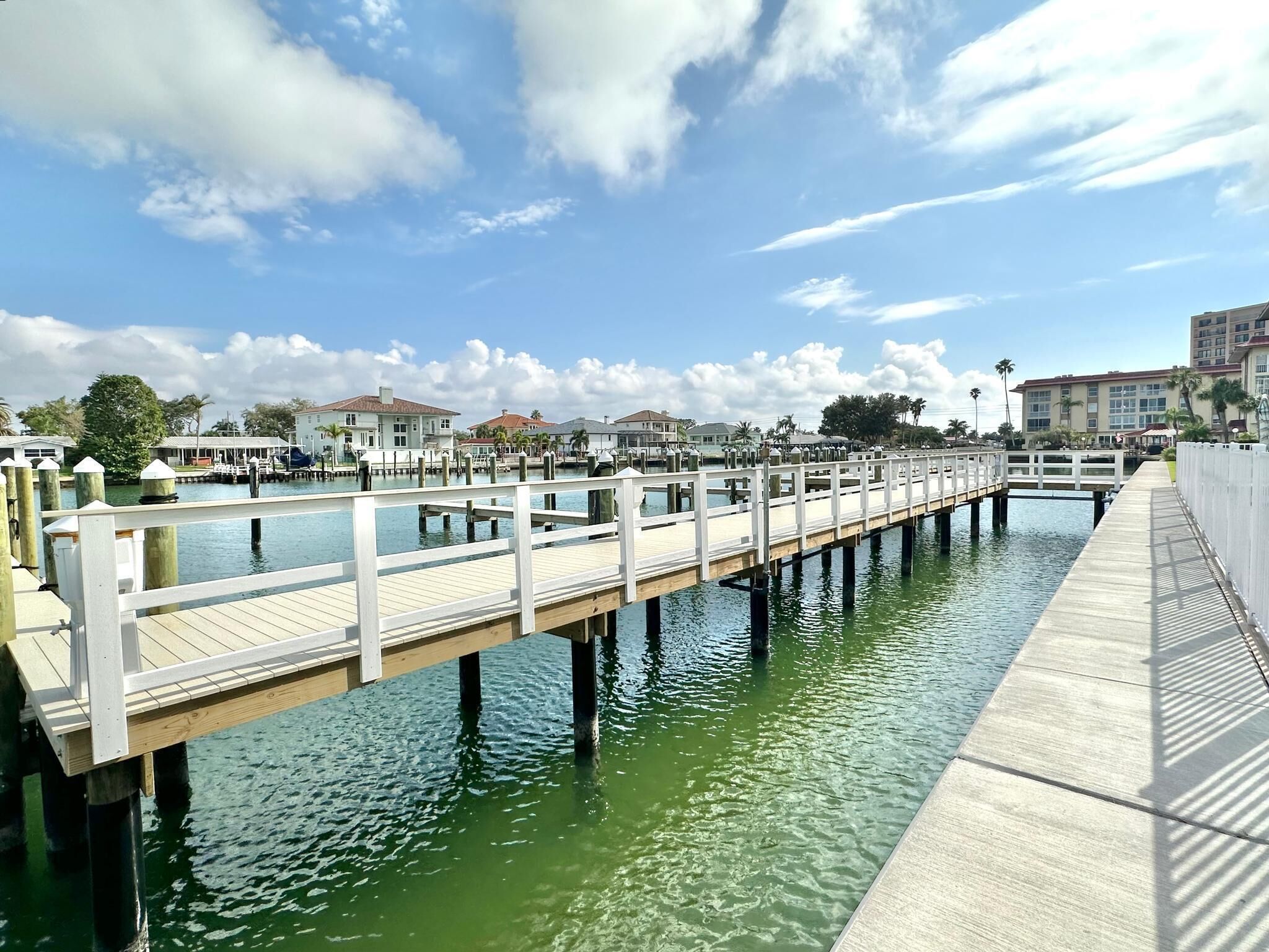 Un muelle frente al agua con una pasarela de madera que se extiende sobre aguas tranquilas, rodeado de edificios y palmeras.