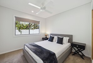 A modern bedroom with a large bed, a ceiling fan, a window with a view of greenery, and a bedside table with a lamp. - Oceanfront View Beachside Bliss – Ocean Views, Plunge Pool & Pet Friendly (Sapphire Beach)