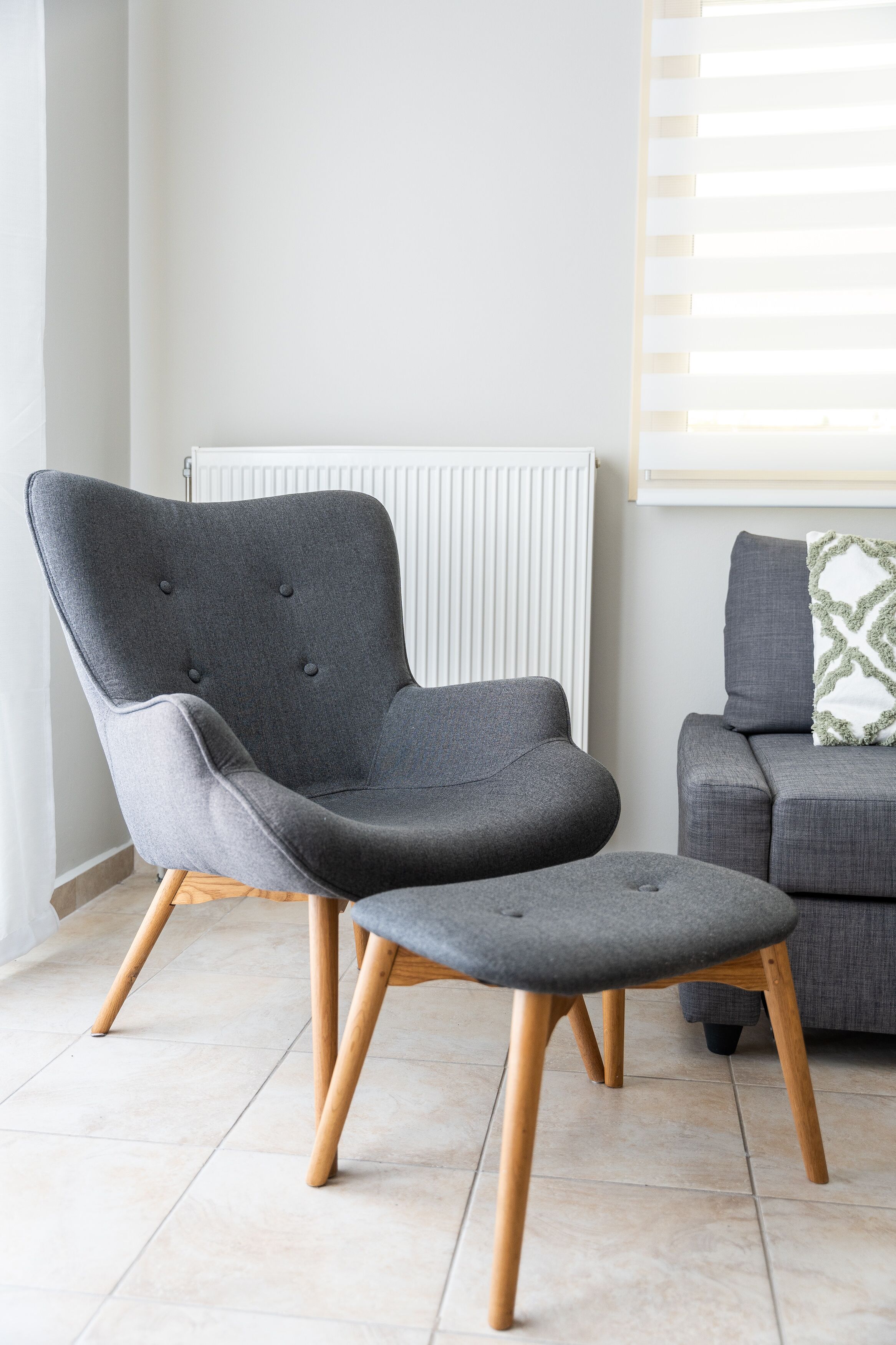 A modern living room with a grey armchair and ottoman, wooden floor, and a grey sofa with a patterned pillow.