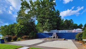 A paved patio area with a grill and seating, surrounded by a blue fence and lush greenery. - Lakefront Two Bedroom Cabin #34 (Charlotte)