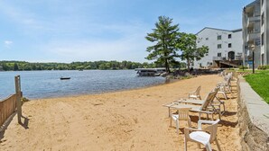A sandy beach with lounge chairs and a wooden fence, a boat on the water, and a building with a balcony. - Some'R on the Beach - Beachfront Condo for Six (Wisconsin Dells)