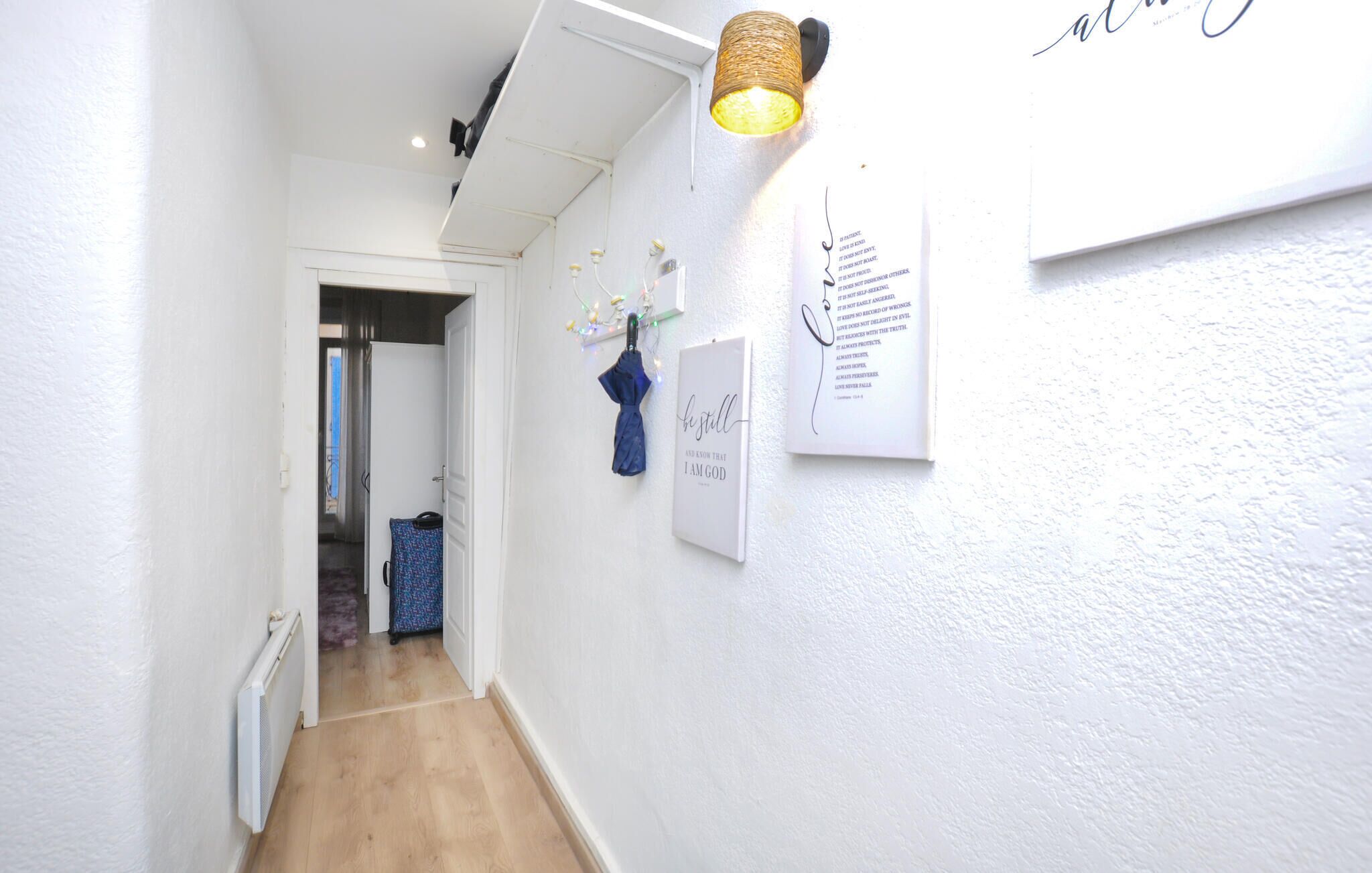 A narrow hallway with white walls, a wooden floor, and a ceiling light fixture. There are two framed inspirational quotes on the wall.