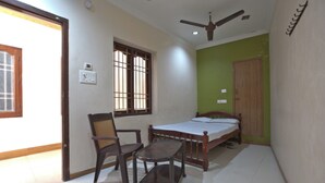 A bedroom with a bed, a chair, a table, a ceiling fan, and a window with bars. - JAYA BHARATHI LODGE (Vilavancode)