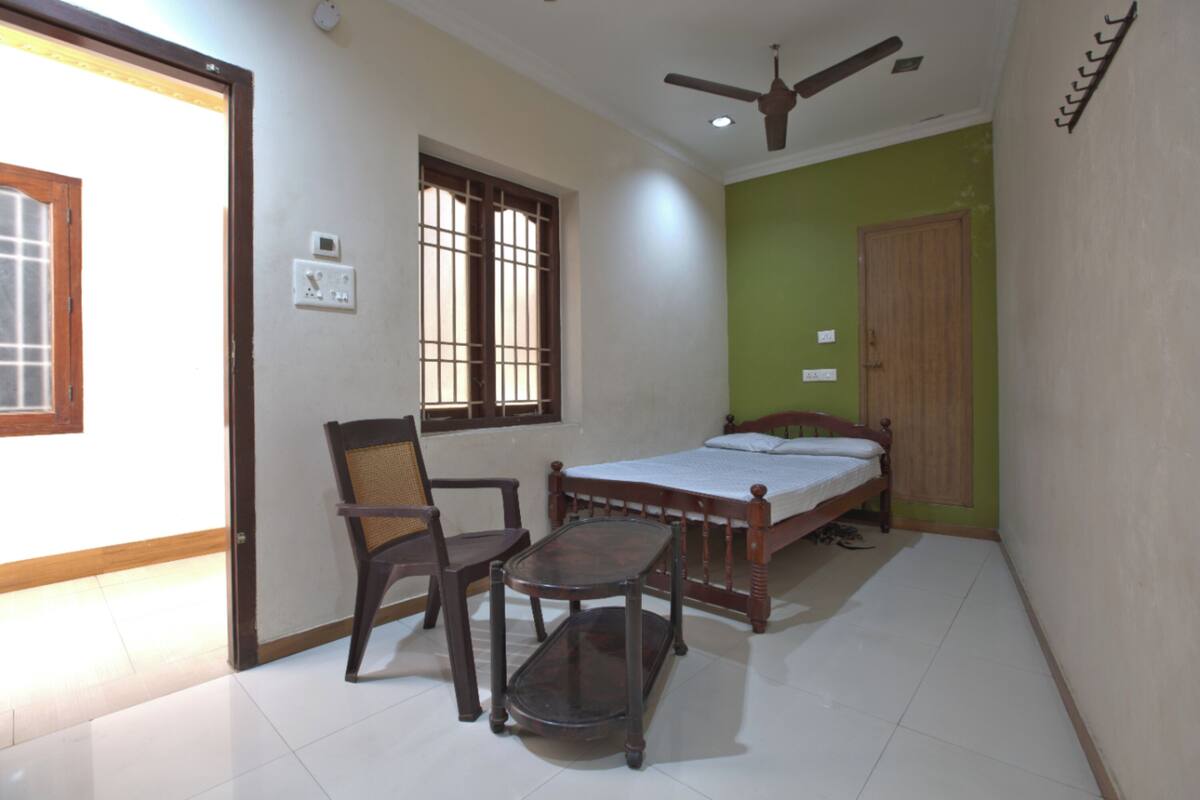 A bedroom with a bed, a chair, a table, a ceiling fan, and a window with bars.