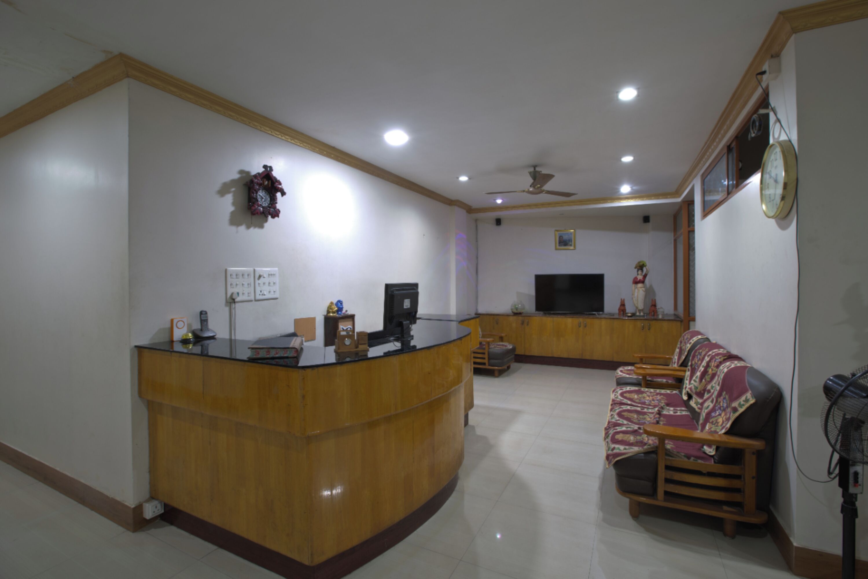 A spacious living area with a wooden reception desk, a sofa, a television, and a ceiling fan.