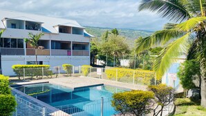 Edificio de dos pisos con piscina, rodeado de palmeras y una zona delimitada por una cerca.