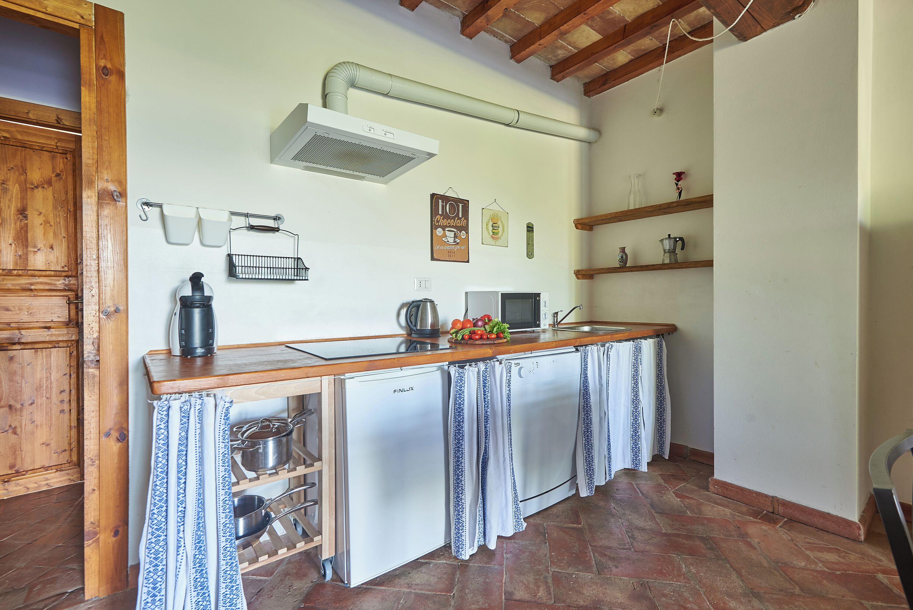 A compact kitchen with a built-in dishwasher, a microwave, and a coffee maker on the counter. There are hanging cups and a small shelf with decorative items.