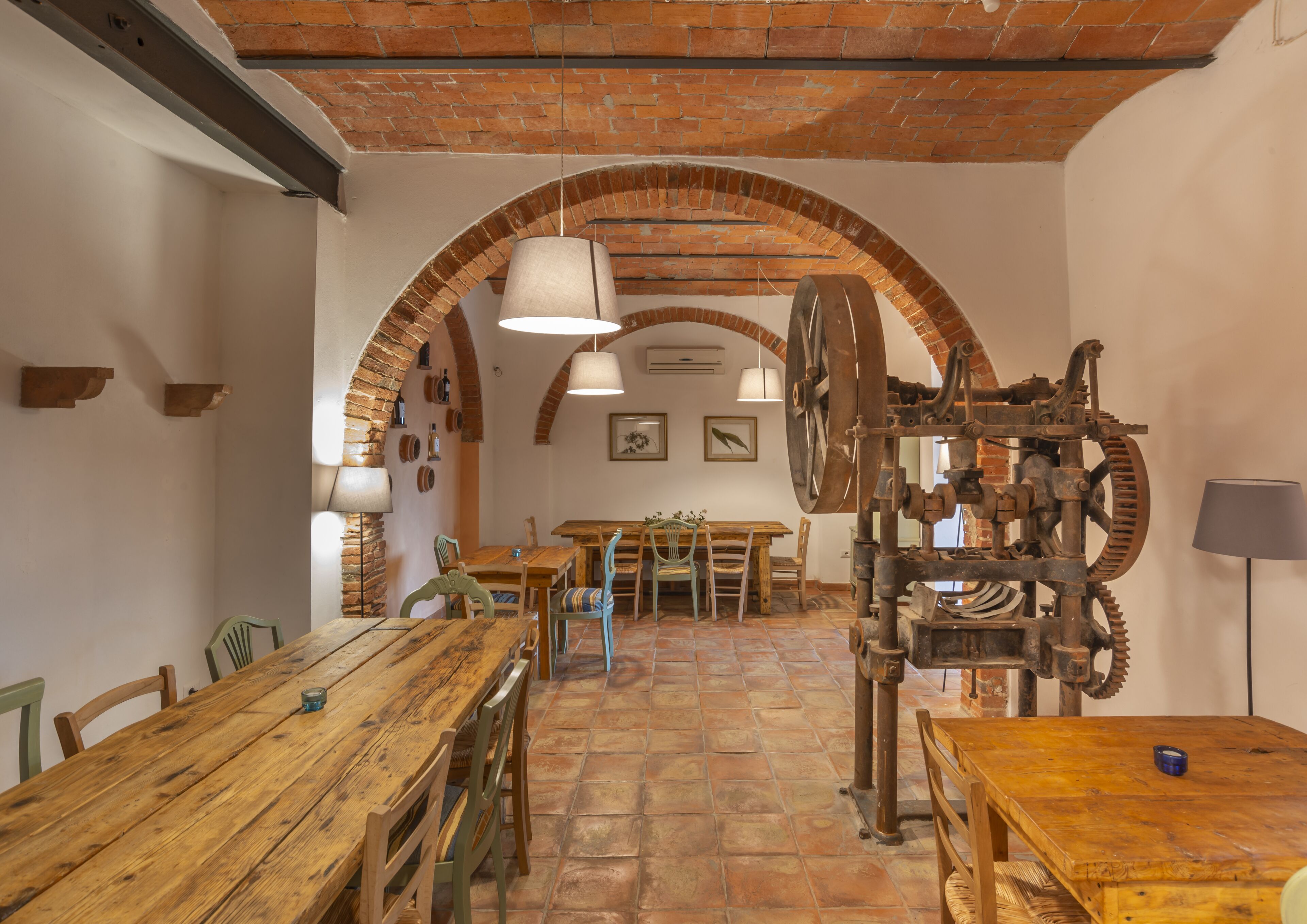 Un'area pranzo rustica con tavoli e sedie in legno, archi in mattoni e una grande struttura a ruota meccanica.