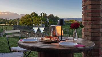 Une table ronde en bois, agrémentée d’assiettes, d’une lampe et d’un vase avec une fleur, est placée à l’extérieur, offrant une vue sur un vignoble et des montagnes en arrière-plan.