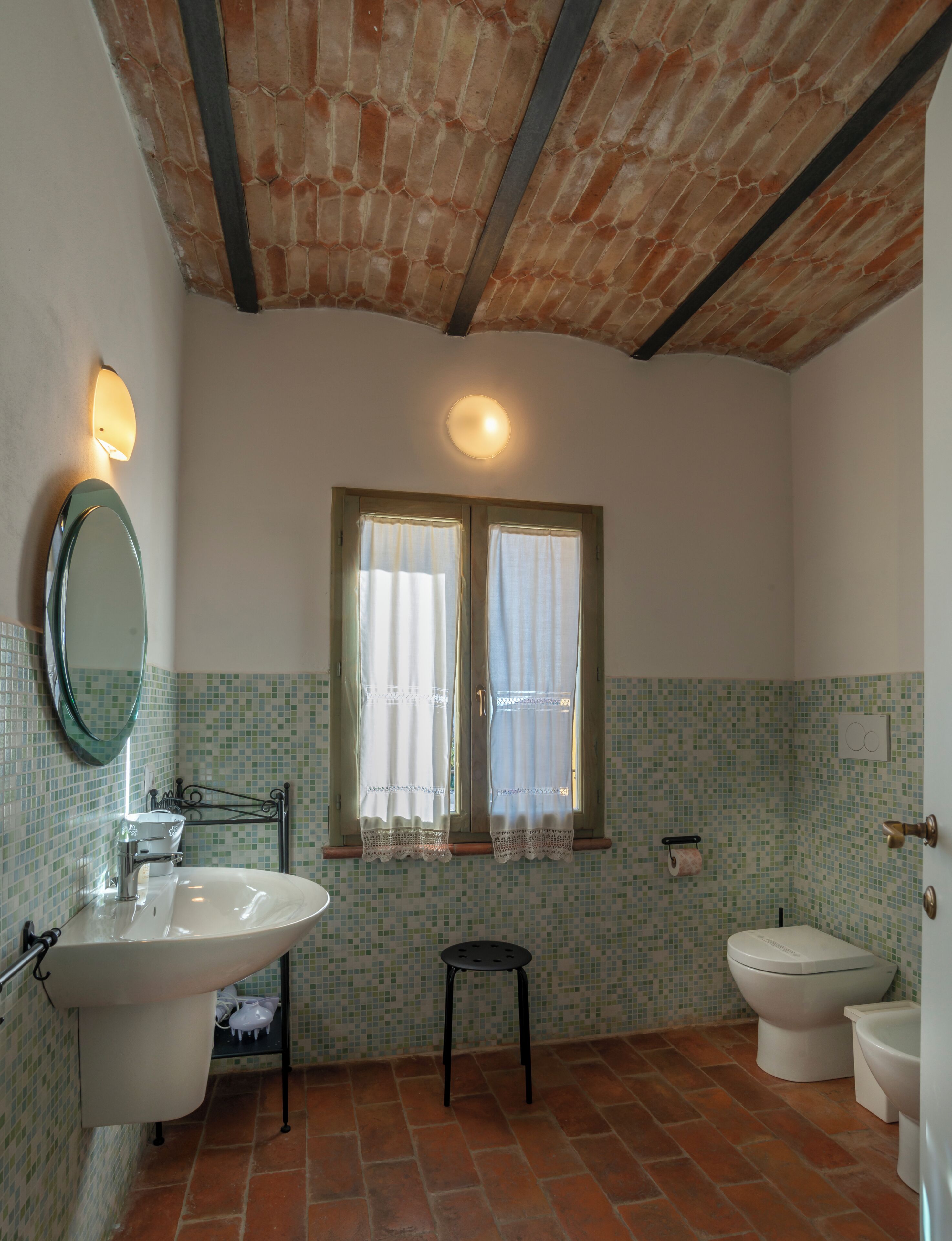A bathroom with a large sink, two toilets, a small stool, a mirror, and a window with curtains.