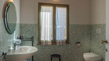 A bathroom with a large sink, two toilets, a small stool, a mirror, and a window with curtains.