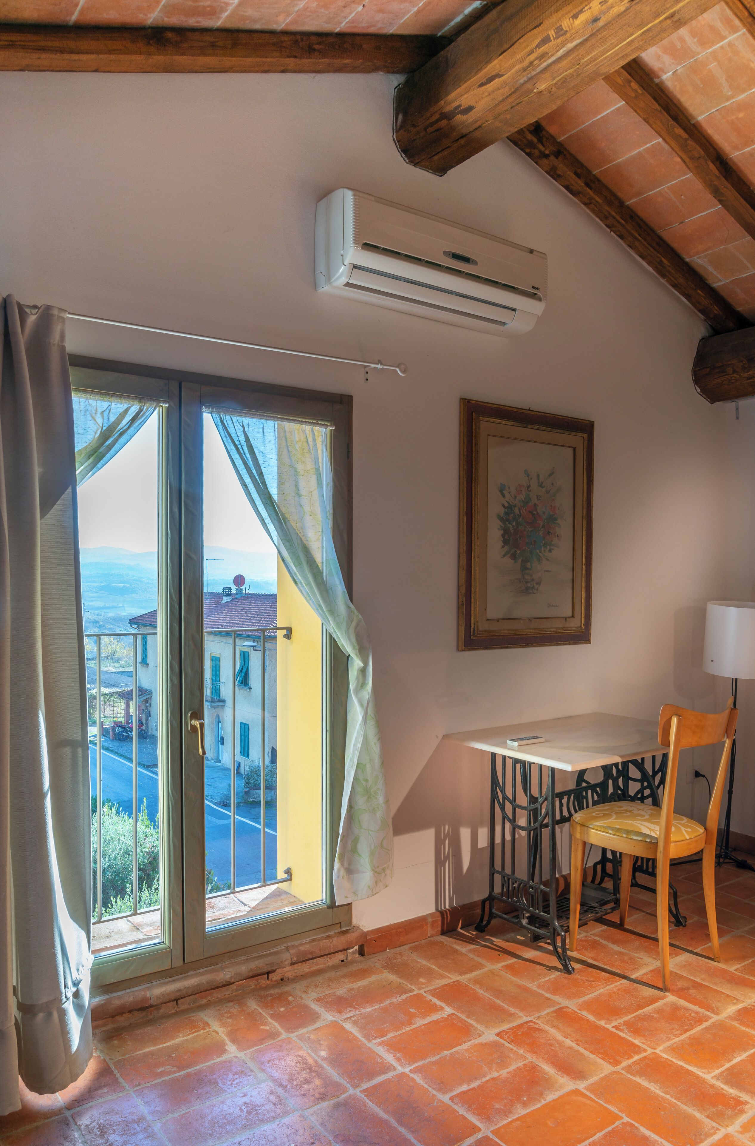 A room with a large window, an air conditioning unit, a table with a chair, and a framed picture on the wall.