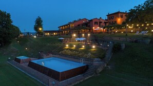 Una piscina grande con una terraza de madera, rodeada por un muro de piedra y un césped. Hay sillas de descanso y sombrillas bajo un porche cubierto.