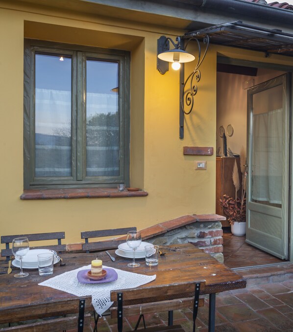 Un área de comedor al aire libre con una mesa de madera preparada para dos personas, una ventana con vistas y una lámpara fijada a la pared.