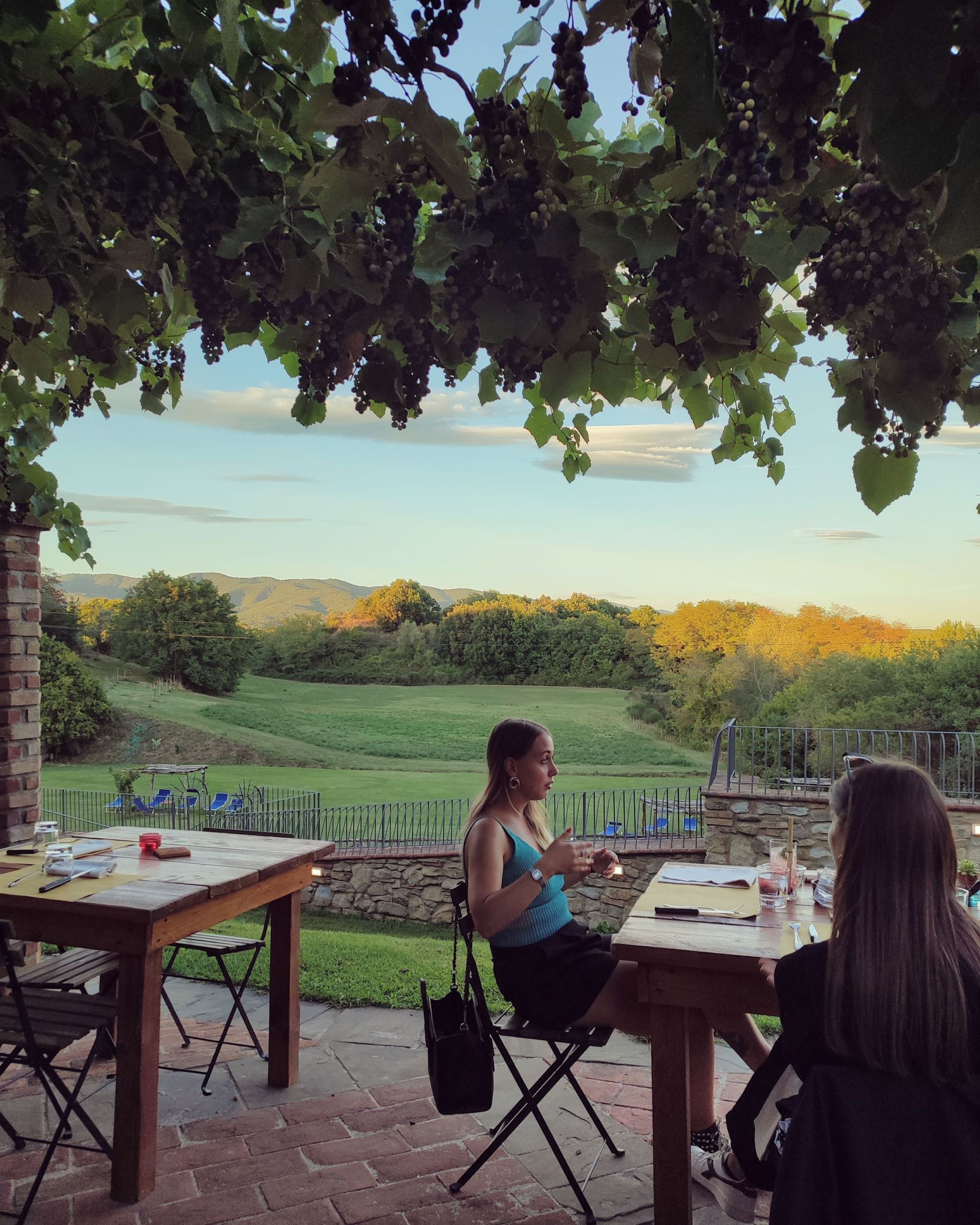 Un área de comedor al aire libre con mesas y sillas, un muro de piedra y vistas a un campo y colinas al fondo.