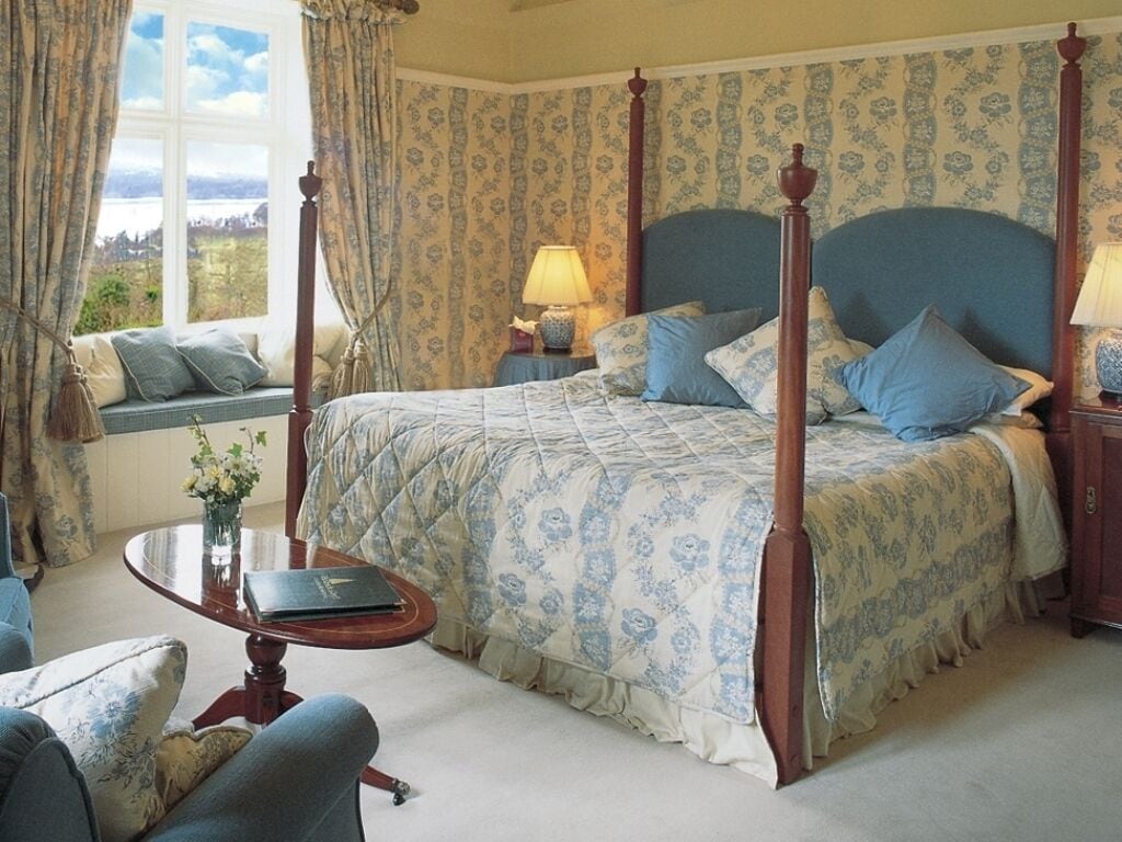 A four-poster bed with a floral bedspread, a bedside table with a lamp, a window seat with a view, and a small table with a book.