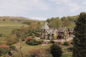 Una casa grande e histórica con un techo empinado, rodeada de jardines exuberantes y un paisaje montañoso.