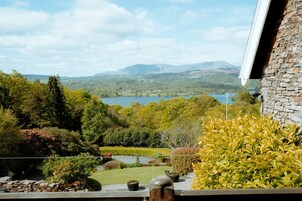 Vista desde un balcón sobre un lago con montañas al fondo, rodeado de exuberante vegetación y un edificio de piedra.