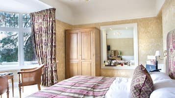 A bedroom with a large bed, a wooden wardrobe, a desk, and a chair. There is a window with curtains, a mirror, and a chandelier.