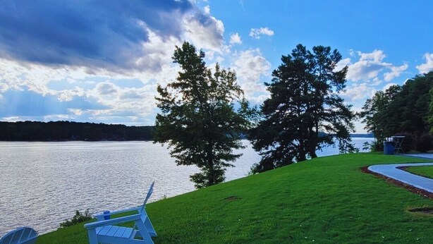 A lakeside view with two Adirondack chairs on a grassy area, overlooking a calm lake and a forested shoreline. - Lakefront Two Bedroom Cabin #35 (Charlotte)