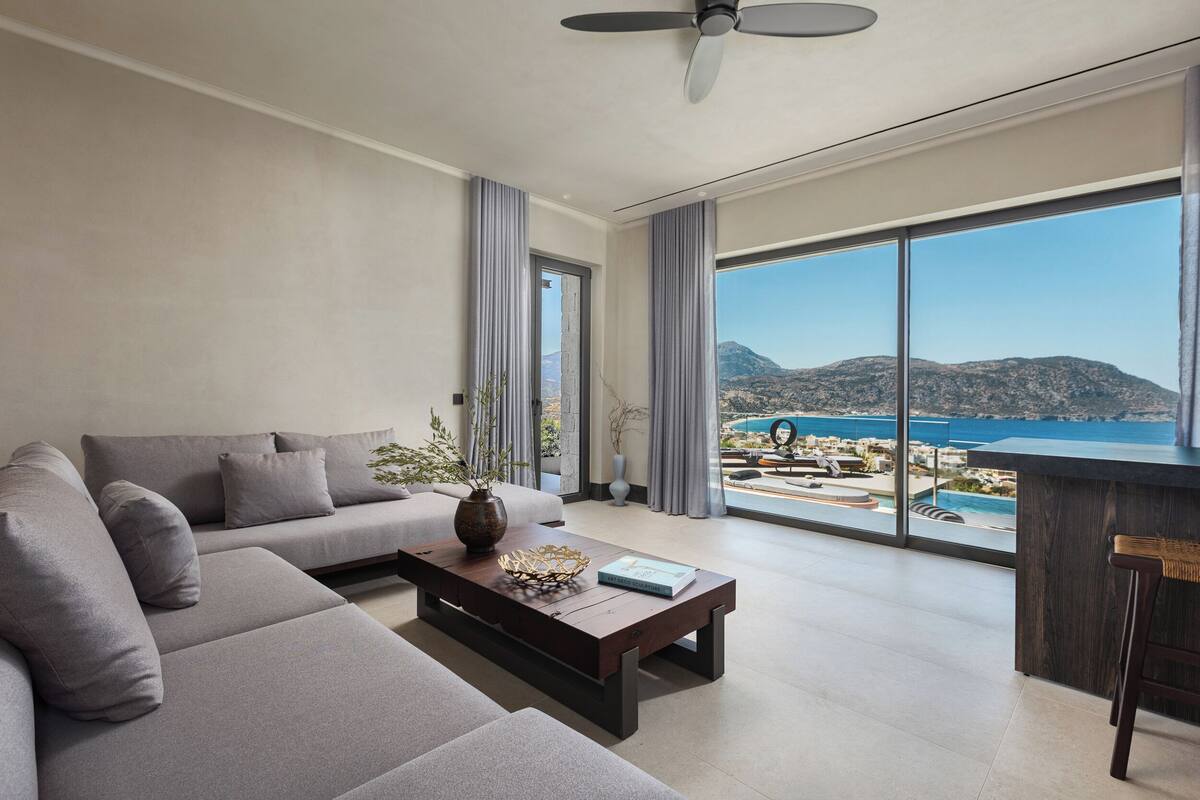 A modern living room with a sofa, coffee table, and large windows offering a view of a pool and mountains.