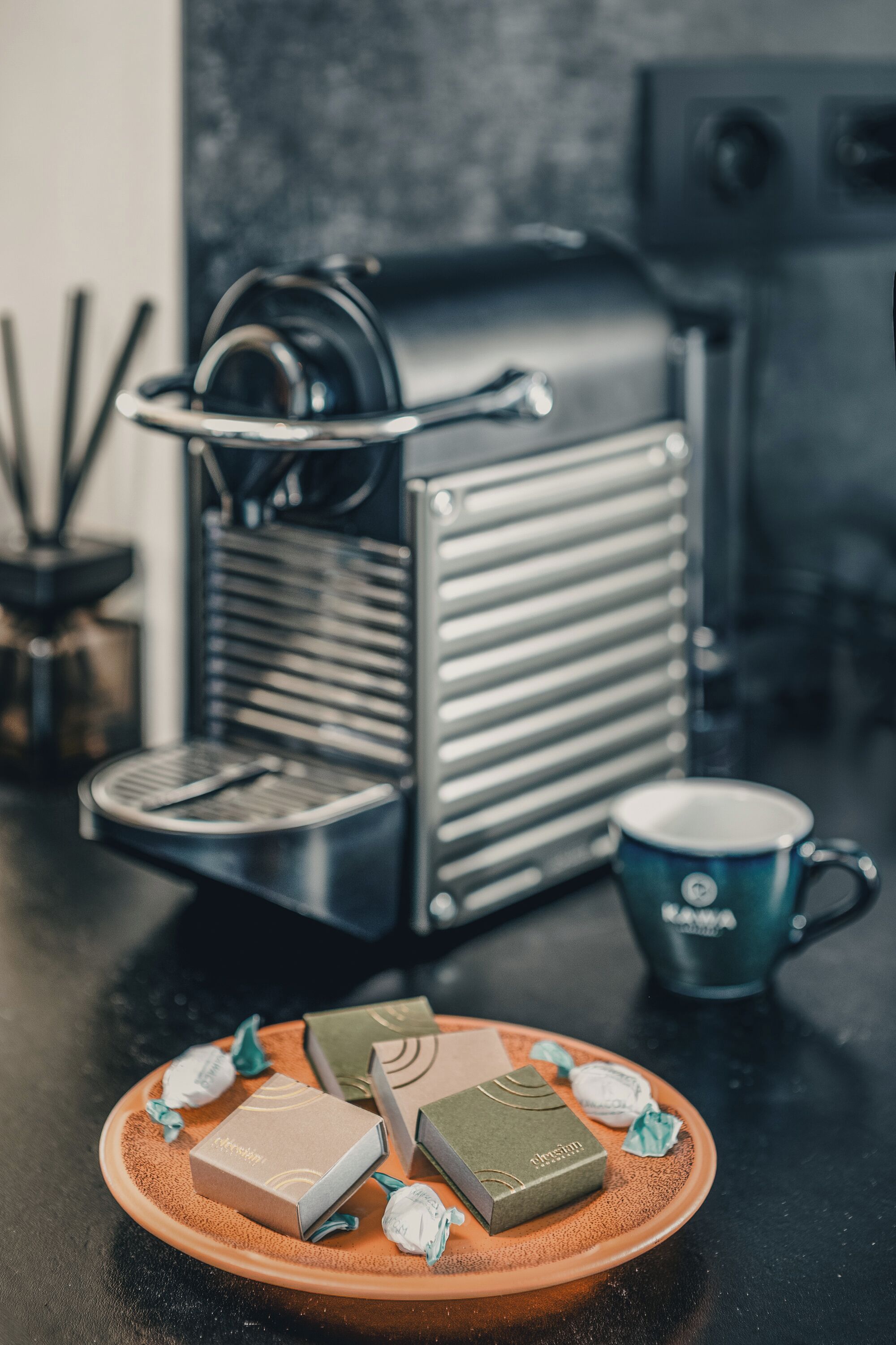 A coffee machine, a cup, and a plate with various packaged items on a table.