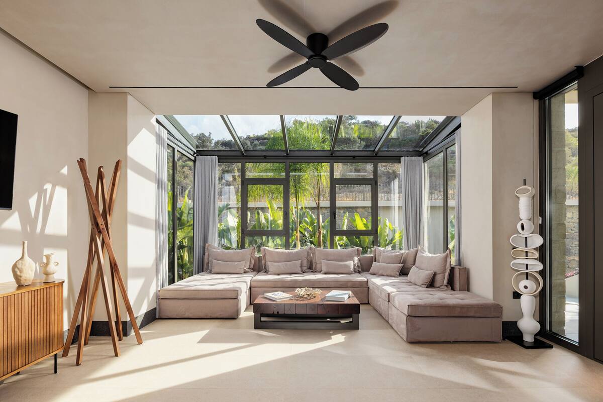 A modern living room with a sectional sofa, a ceiling fan, and large windows offering a view of greenery.