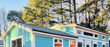 Una casa pequeña de un solo piso con fachada azul y detalles blancos, que cuenta con un porche con dos sillas Adirondack y una escalera que lleva a una terraza.