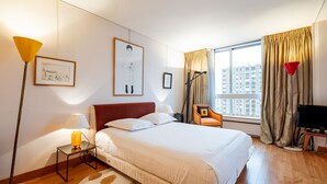 A hotel room with a large bed, a bedside table with a lamp, a chair, a TV, and a window with curtains.