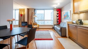 A modern living room with a dining area, a sofa, a TV, and a kitchen with wooden cabinets and a window with a city view.