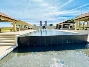 Ein Swimmingpool mit schwarzem Rand, umgeben von überdachten Sitzbereichen und blauem Himmel.