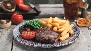 Une assiette contenant un steak, des frites, des petits pois et une tomate grillée, le tout posé sur une table en bois rustique.