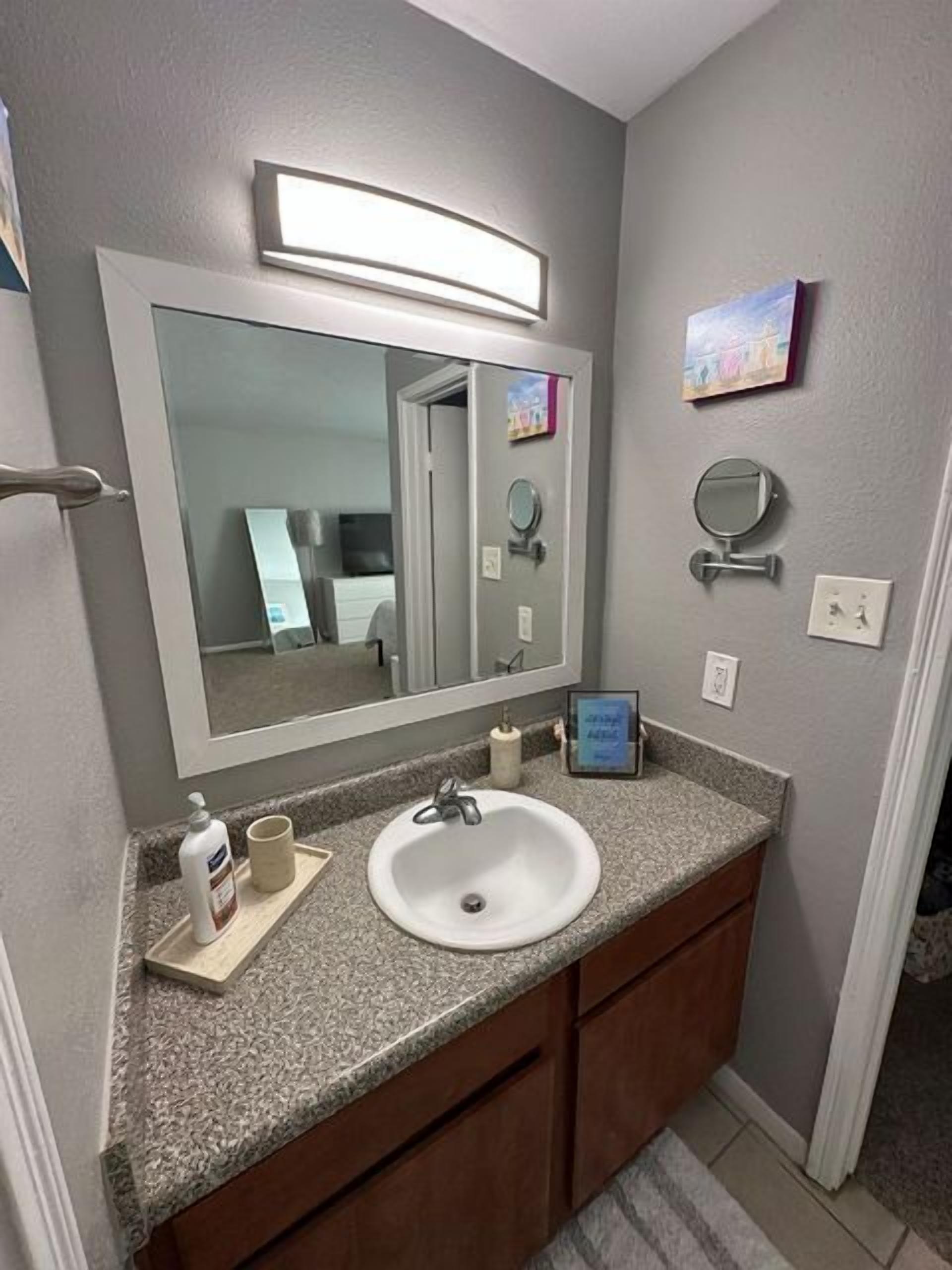 Un baño con un lavabo blanco, un espejo grande, un tocador con encimera y un pequeño espejo redondo en la pared.
