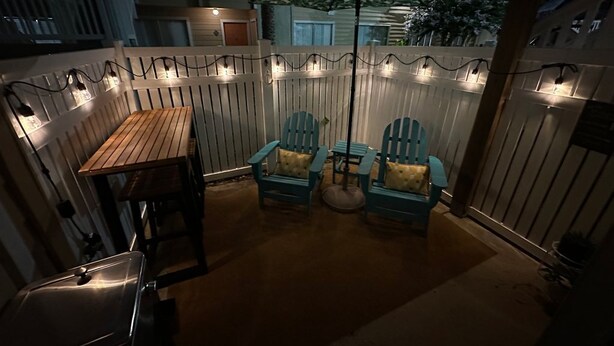 En udendørs terrasse med et træbord og to blå stole, omgivet af et træhegn og lyskæder.