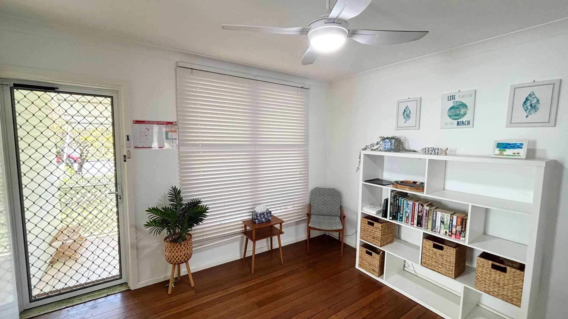Una habitación con ventilador de techo, una estantería, una silla, una mesita pequeña y una planta en maceta.