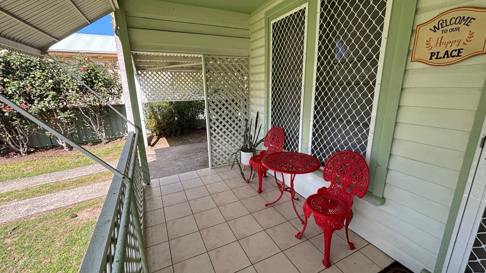 A covered porch with a 'Welcome to Our Happy Place' sign, red metal chairs, and a potted plant.